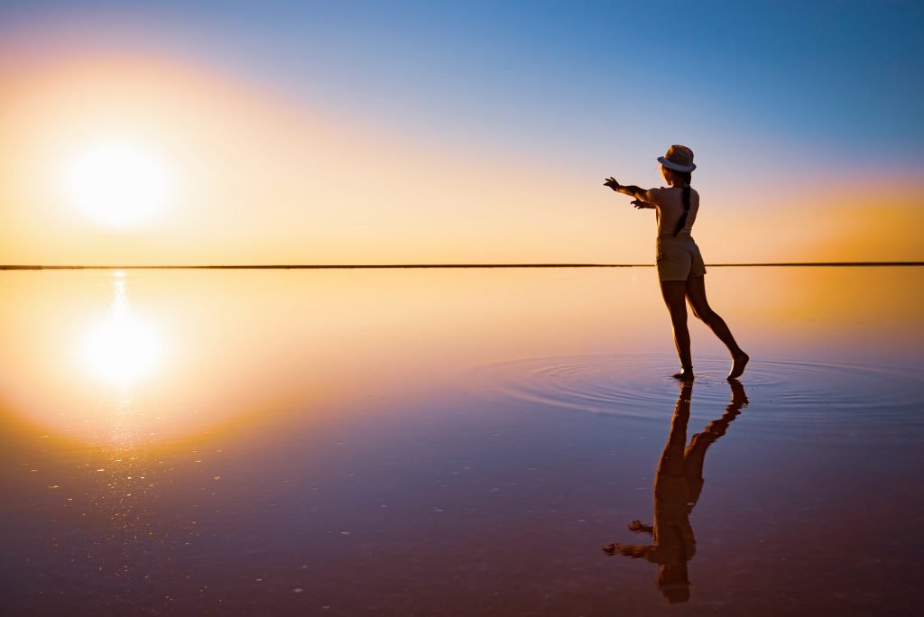 A girl reaches out with her hands to the setting sun on the mirror surface of a salt lake